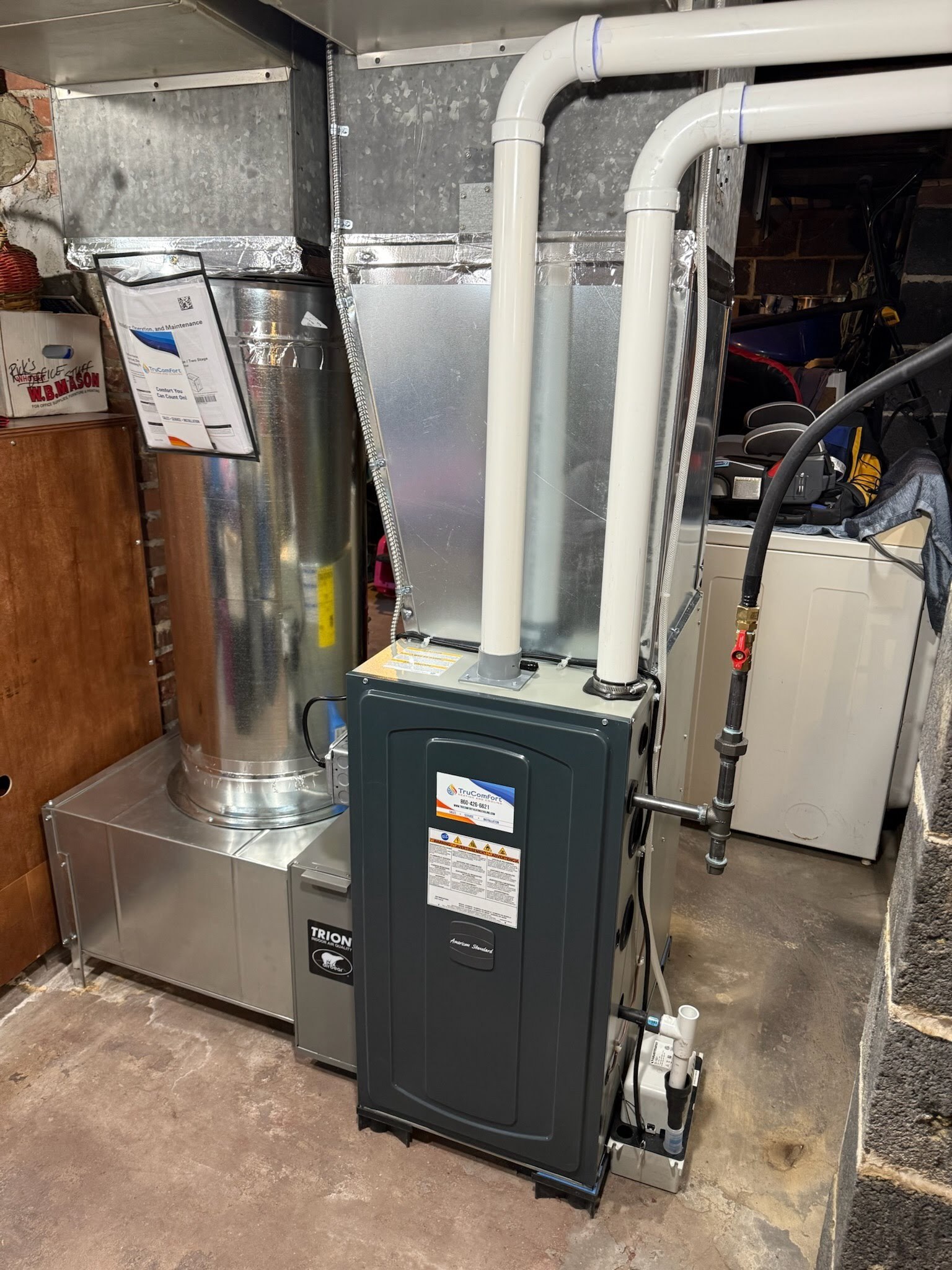 A residential HVAC unit neatly installed in a basement, featuring metal ductwork, PVC exhaust and intake pipes, a condensate pump, and electrical wiring. The equipment includes clearly labeled maintenance documentation. The surrounding basement area has concrete floors and storage items.